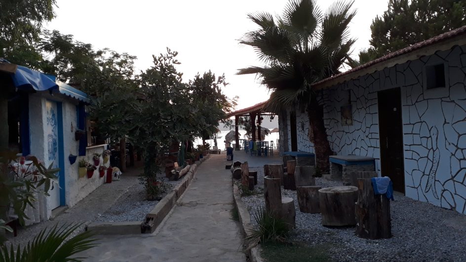 Bir plaj restoranının gölgelikli terasından Zeytinlikuyu koyuna bakış. Ön planda masada pembe çiçekler, arka planda ise kumsaldaki hasır şem