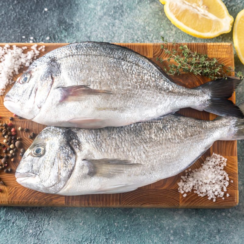 Ahşap bir kesme tahtası üzerinde yan yana duran iki taze çipura balığı. Balıkların etrafında deniz tuzu, tane karabiber, taze kekik dalları 